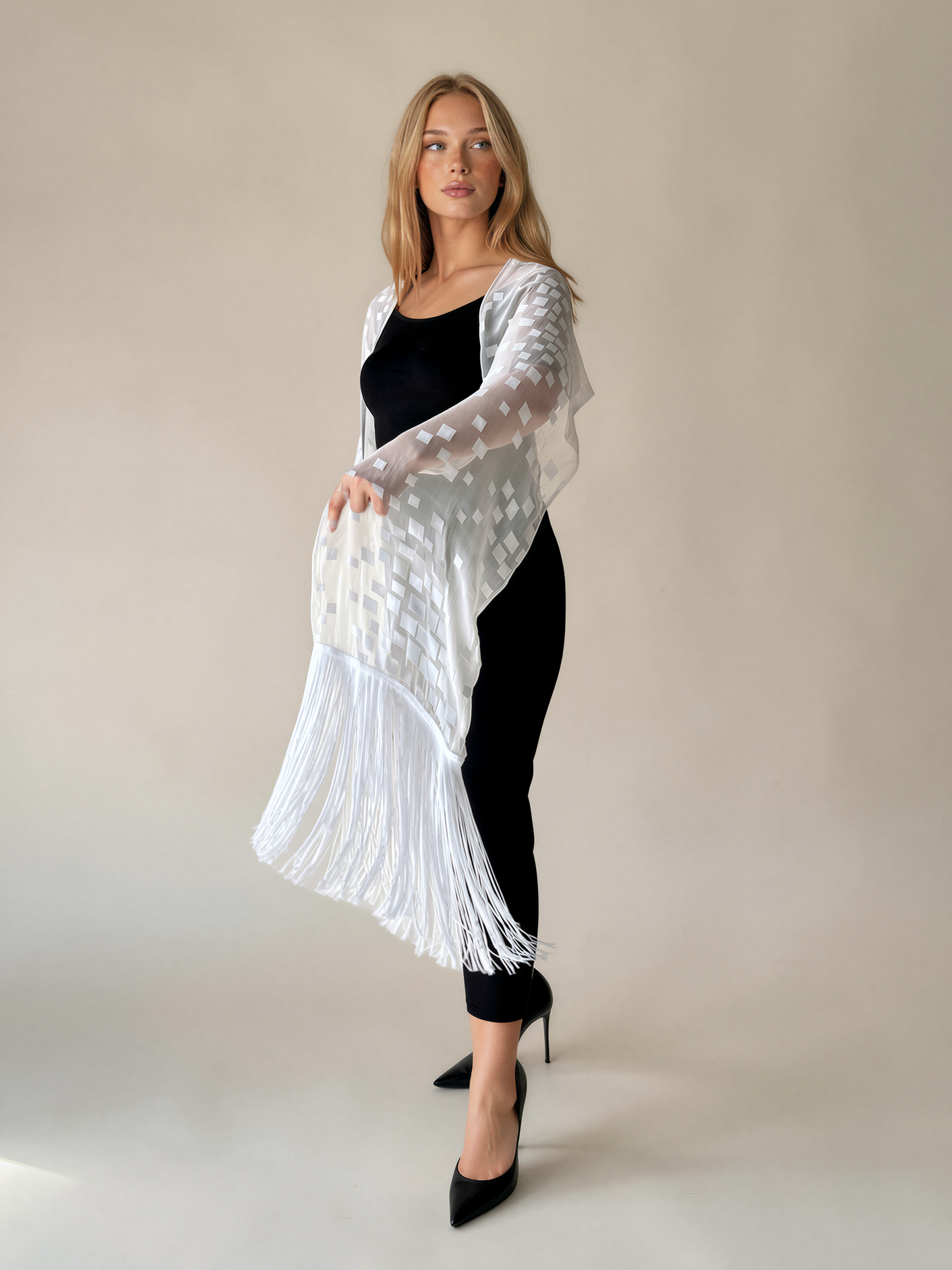 A woman models a white sheer long-sleeve cardigan with a textured pattern and fringe hem, worn over a black top and black pants against a neutral beige studio background.