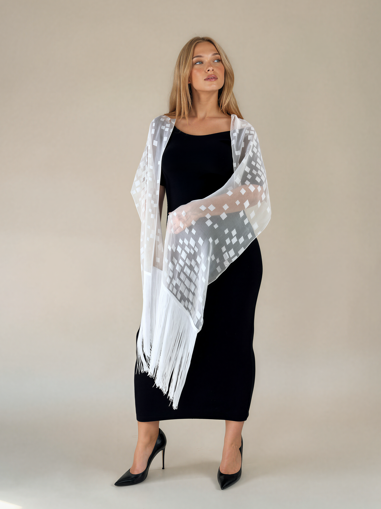 A woman modeling a white sheer patterned shawl with fringe over a black midi dress and black pointed heels, standing against a plain beige studio background.