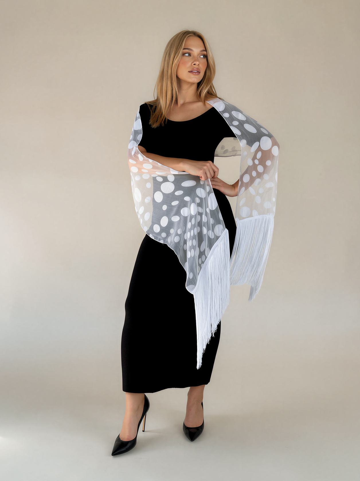 A woman modeling a white fringed shawl with gray and peach polka dots, worn over a black dress against a plain beige studio background.