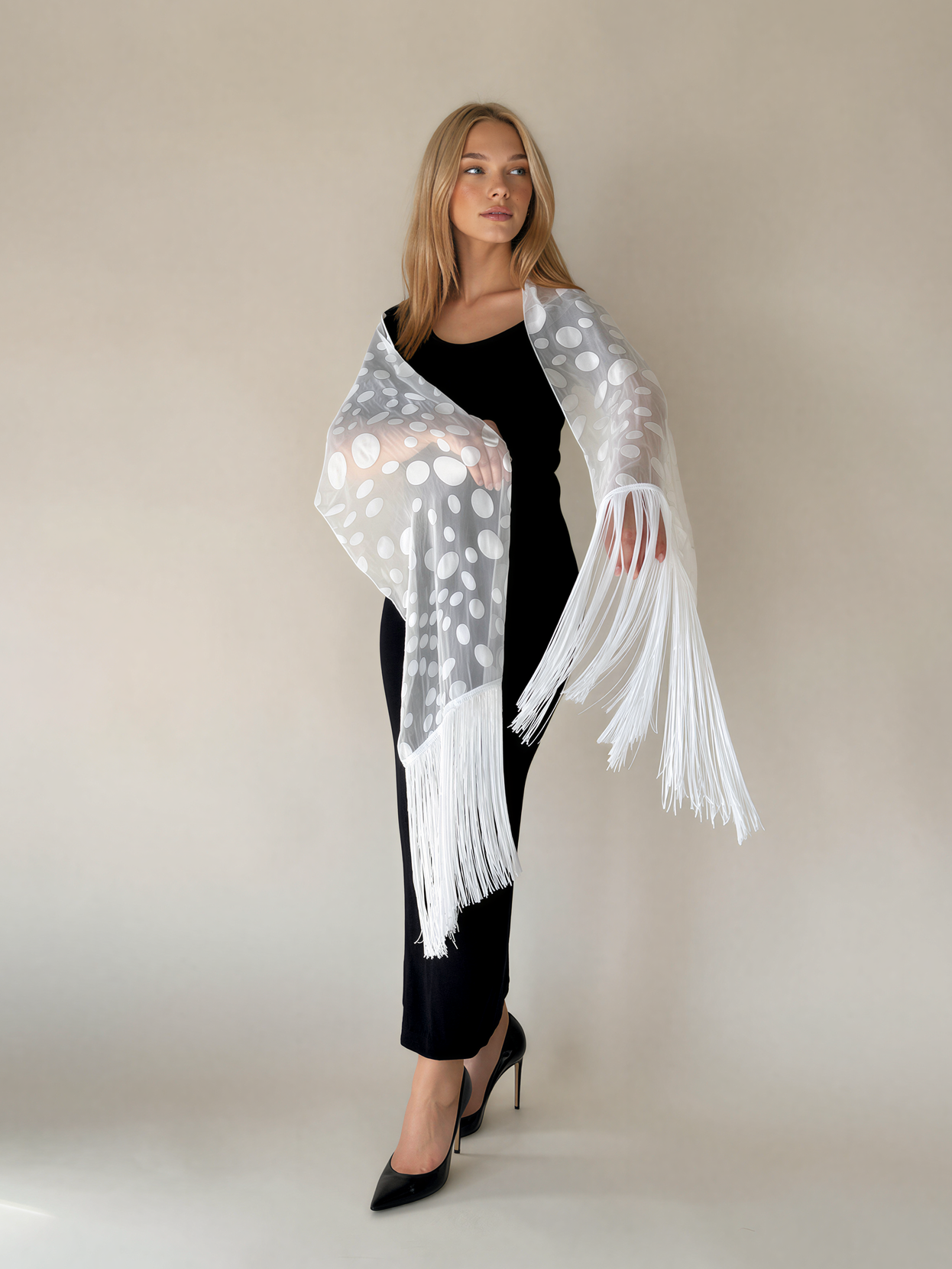 A woman modeling a sheer white shawl with silver polka dots and long white fringe, worn over a black outfit against a plain beige studio background.