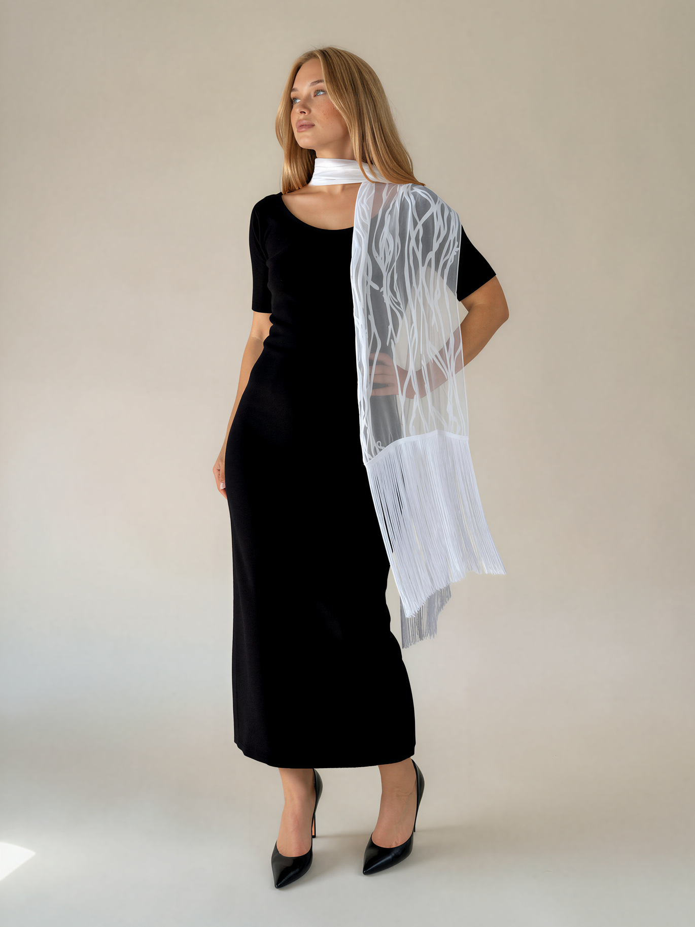 A woman modeling a black midi dress with a white sheer overlay draped over one shoulder, set against a plain beige studio background.