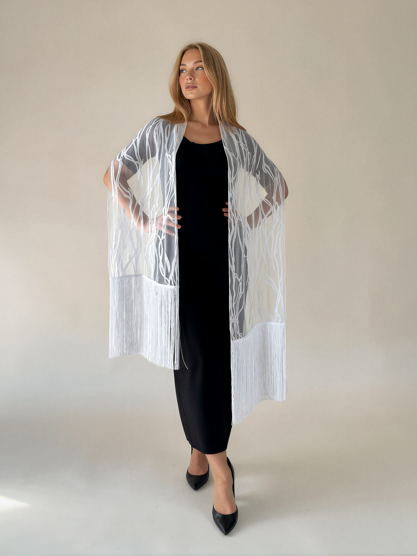A woman modeling a sheer white open-front cardigan with fringe details over a black dress, standing against a plain beige studio background.
