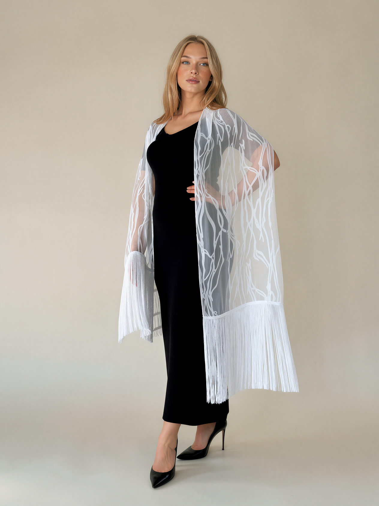 A woman modeling a white open-front fringed cardigan or wrap with abstract leaf-like embroidery over a black dress, standing against a plain beige studio background.