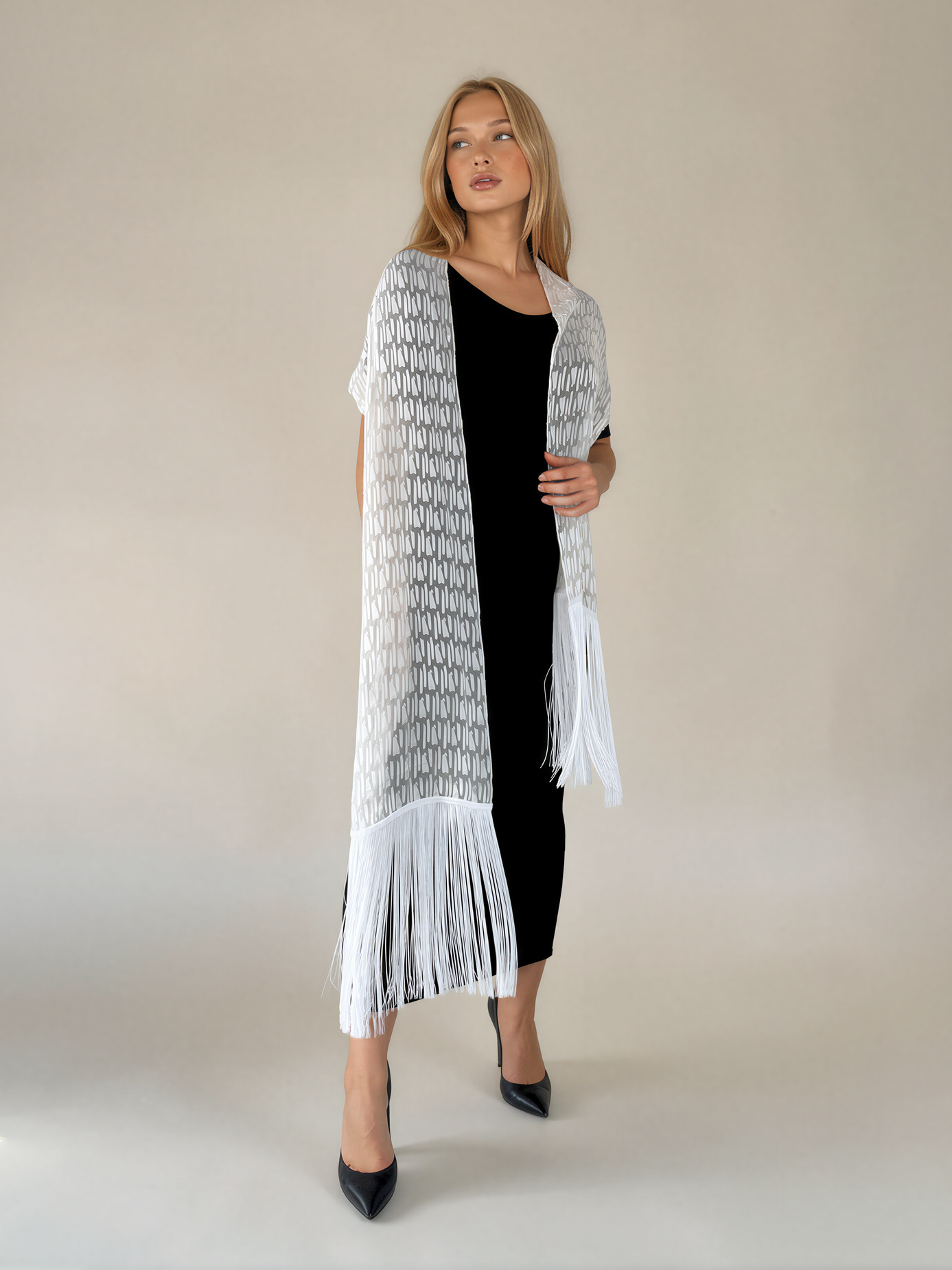 A woman modeling a long white knit cardigan with a black geometric pattern and long fringe, worn open over a black dress, against a plain beige studio background.