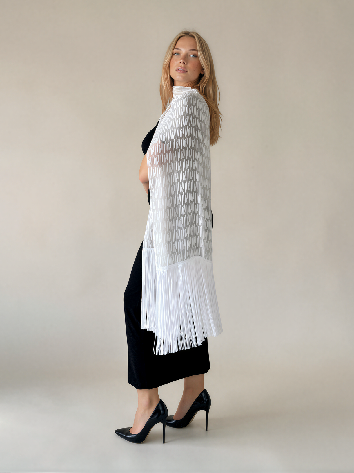 A woman stands in profile wearing a white open-knit fringed shawl or wrap over a black dress, against a plain light beige studio background.
